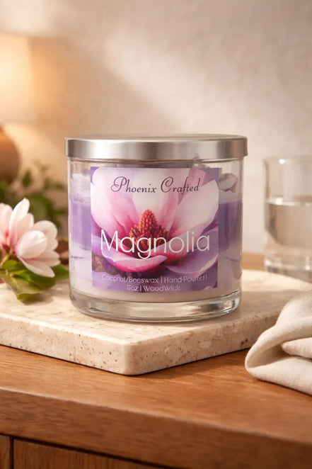 Clear glass jar candle with silver lid and purple layered wax; label reads Phoenix Crafted and Magnolia with magnolia artwork on a marble tile.