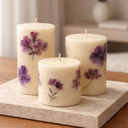 Three cream pillar candles with purple flower designs and unlit wicks on a rectangular stone slab atop a wooden table