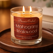 Clear glass jar candle labeled 'Phoenix Crafted Mahogany Teakwood' with two lit wicks and creamy wax on a round wooden tray, warm lamp-lit background