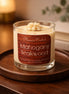 Glass jar candle with cream-colored wax and piped topper labeled "Phoenix Crafted Mahogany Teakwood" on a round wooden tray.