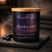 Black glass candle jar with light wooden lid and purple label 'Phoenix Crafted - Obsidian Mist' on textured slate with black stones and smoke.