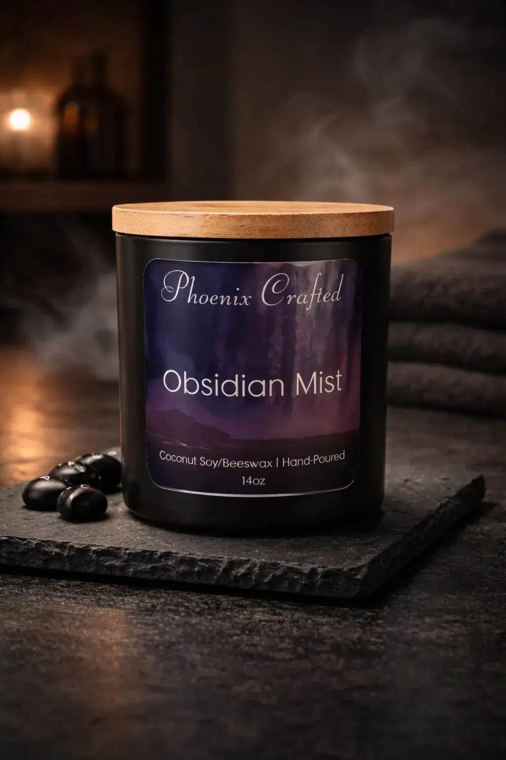 Black glass candle jar with light wooden lid and purple label 'Phoenix Crafted - Obsidian Mist' on textured slate with black stones and smoke.