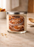 Clear glass jar candle with silver lid and pale cream wax; front label reads "Peach Cobbler" showing a cobbler image.