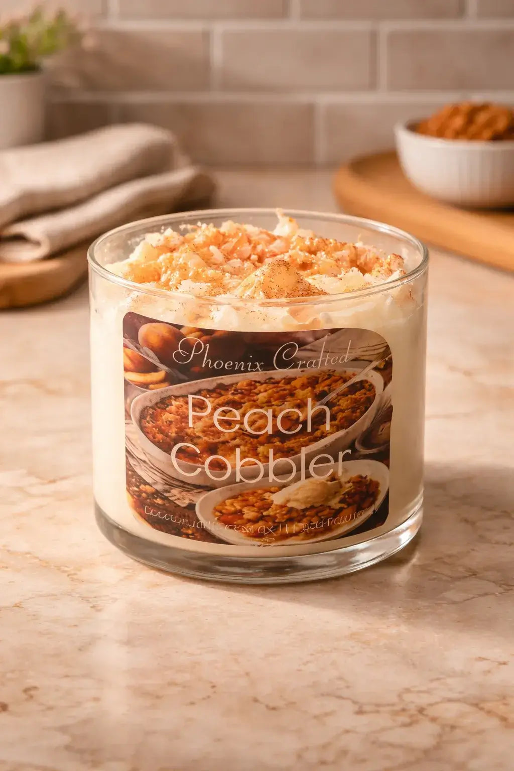 Clear round glass candle with white whipped wax and peach crumble topping labeled 'Phoenix Crafted' and 'Peach Cobbler'.