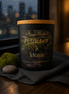 Black candle jar with tan lid and gold "Phoenix Crafted Petrichor & Moss" label over a moss image, beside a small moss ball and smooth gray stone.