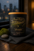 Black candle jar with tan lid and gold "Phoenix Crafted Petrichor & Moss" label over a moss image, beside a small moss ball and smooth gray stone.