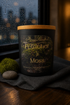 Black candle jar with tan lid and gold "Phoenix Crafted Petrichor & Moss" label over a moss image, beside a small moss ball and smooth gray stone.