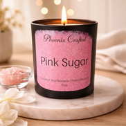 Black matte jar candle with a pink 'Phoenix Crafted' label on a round coaster beside a bowl of pink granules and white flowers.