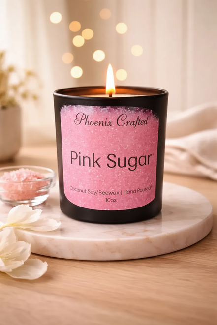 Black matte jar candle with a pink 'Phoenix Crafted' label on a round coaster beside a bowl of pink granules and white flowers.