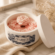 Round white lidded candle tin with pink sculpted wax roses, central wooden wick, blue floral label, on beige cloth and marble tray with glass bottle.