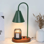 Deep green arched candle warmer lamp with a conical shade above a textured jar candle, styled beside a white vase of dried stems.