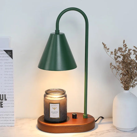 Deep green arched candle warmer lamp with a conical shade above a textured jar candle, styled beside a white vase of dried stems.