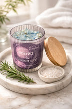Sea Salt & Rosemary Candle in a glass jar with a wooden lid on a marble tray alongside fresh rosemary and sea salt.