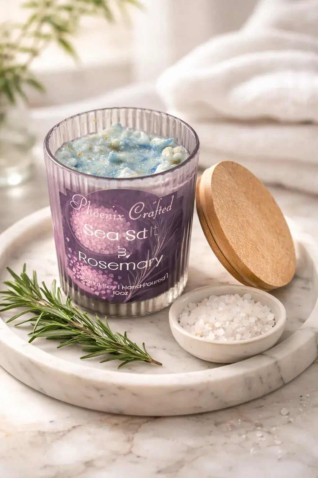 Sea Salt & Rosemary Candle in a glass jar with a wooden lid on a marble tray alongside fresh rosemary and sea salt.