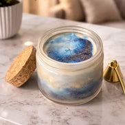Sea Salt & Rosemary Candle in a decorative jar with blue and gold hues on a marble surface.