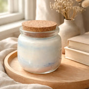 Sea Salt & Rosemary Candle in glass jar with cork lid, displayed on wooden tray near books and dried flowers.