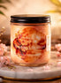 Clear glass jar candle with black screw lid and peach-pink wax labeled Phoenix Crafted - Sparkling Blossom on a round marble slab with pink blossoms.