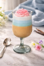 A colorful layered dessert in a glass with pink whipped cream on top, surrounded by flowers and a spoon.