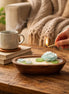 Teak & Mint Renewal Decorative Candles with floral designs and a hand lighting a wick on a cozy wooden table.