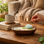 Teak & Mint Renewal Decorative Candles with floral designs and a hand lighting a wick on a cozy wooden table.