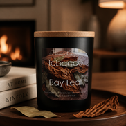 Matte black jar candle with wooden lid labeled 'Tobacco & Bay Leaf' sits on a wooden tray with bay and dried tobacco leaves.