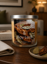 Jar candle with lid and label on a table beside a folded beige napkin and small dish of bay leaves, blurred lamp behind.