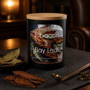 Black matte jar candle with wooden lid and label, dried leaves and small dish to its left, and a brass wick trimmer on a dark placemat.