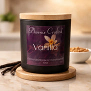 Matte black jar candle with light wood lid on wood coaster, purple label Phoenix Crafted - Vanilla, vanilla pods at left.