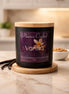 Matte black jar candle with light wood lid on wood coaster, purple label Phoenix Crafted - Vanilla, vanilla pods at left.