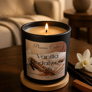 Lit cream wax candle in a matte black jar on a round wooden coaster on a dark wood table, with a vanilla flower and bean pods at right.
