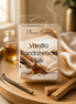 Clear clamshell pack with cream label on a round light-wood tray, beside a vanilla bean, two wooden blocks, amber bottles and cream fabric.