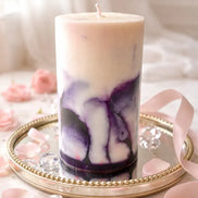 Cylindrical pillar candle with cream top and purple marbled lower half, single wick, on a round mirrored tray with beaded rim, pink petals and ribbon