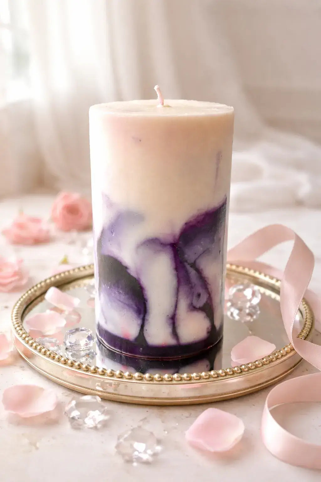 Cylindrical pillar candle with cream top and purple marbled lower half, single wick, on a round mirrored tray with beaded rim, pink petals and ribbon