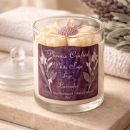 Clear glass jar candle with cream flower-shaped wax top and purple textured center, purple label, and lavender sprigs on a beige stone slab.