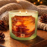 Pale green frosted glass jar candle with two lit wicks and orange label reading 'Phoenix Crafted — Winter Solstice Glow'.