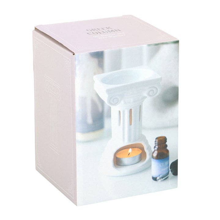White Greek-column ceramic burner with a lit tealight in its base, amber glass bottle with blue label and black cap beside, shown on a pale pink box.