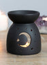 Black oil burner/wax warmer with bowl top and crescent moon and star cutouts showing a tealight, with candles and an amethyst cluster behind.