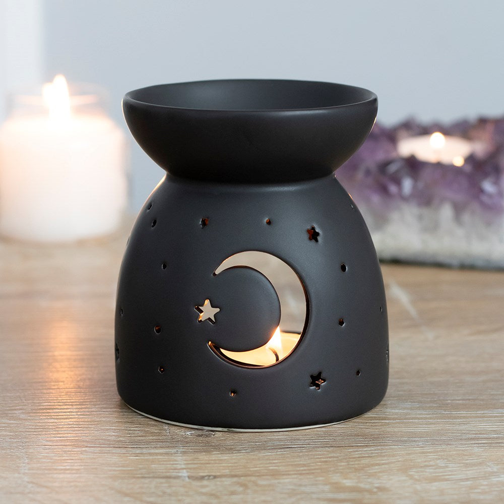 Black oil burner/wax warmer with bowl top and crescent moon and star cutouts showing a tealight, with candles and an amethyst cluster behind.