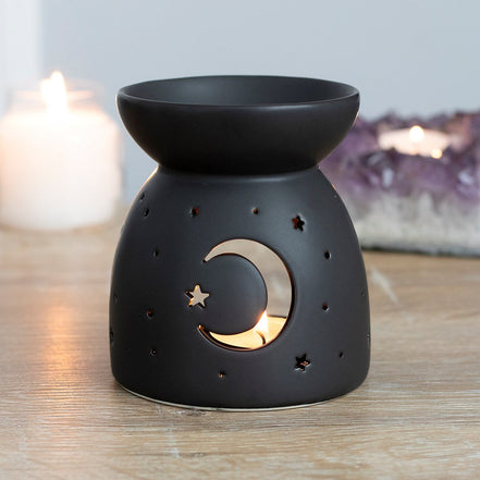 Black oil burner/wax warmer with bowl top and crescent moon and star cutouts showing a tealight, with candles and an amethyst cluster behind.