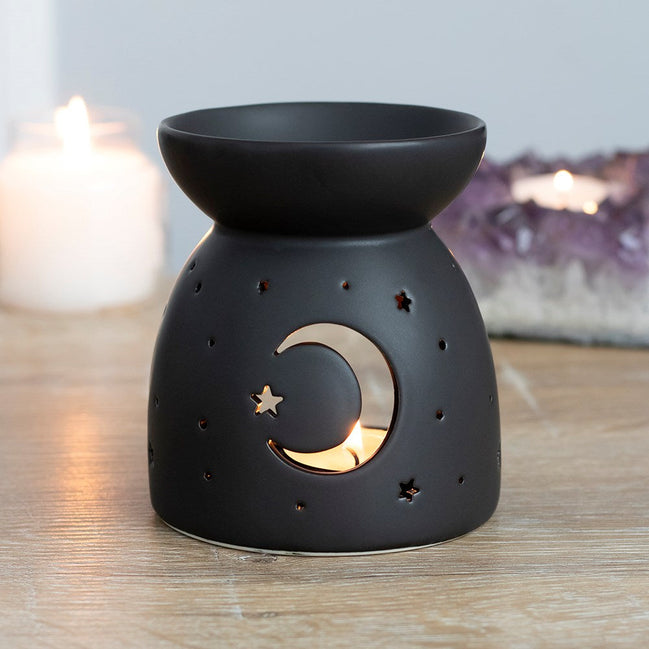 Black oil burner/wax warmer with bowl top and crescent moon and star cutouts showing a tealight, with candles and an amethyst cluster behind.