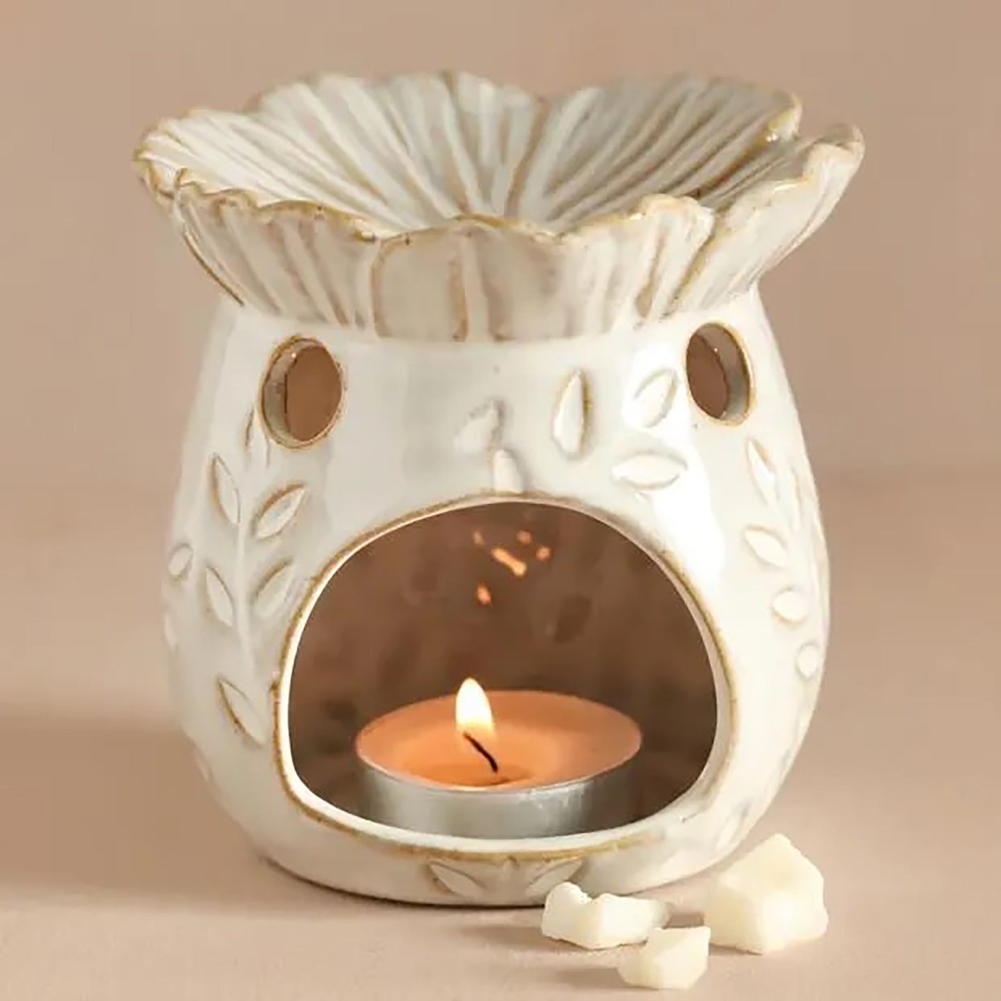 Cream ceramic wax warmer with scalloped bowl, gold trim, leaf pattern, round vents and arched front revealing a lit tealight in a metal cup and wax.