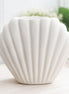 Seashell-shaped matte off-white ceramic vessel with vertical scalloped ridges and open top, on a light wood table with blurred greenery behind.