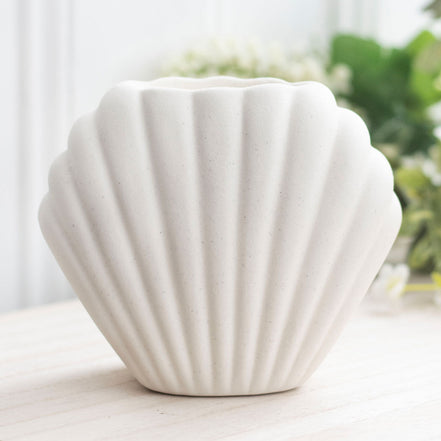 Seashell-shaped matte off-white ceramic vessel with vertical scalloped ridges and open top, on a light wood table with blurred greenery behind.
