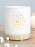 White cylindrical ceramic wax warmer with mandala cutout glowing on a light wood-effect base, beside a blurred candle and small white flowers.