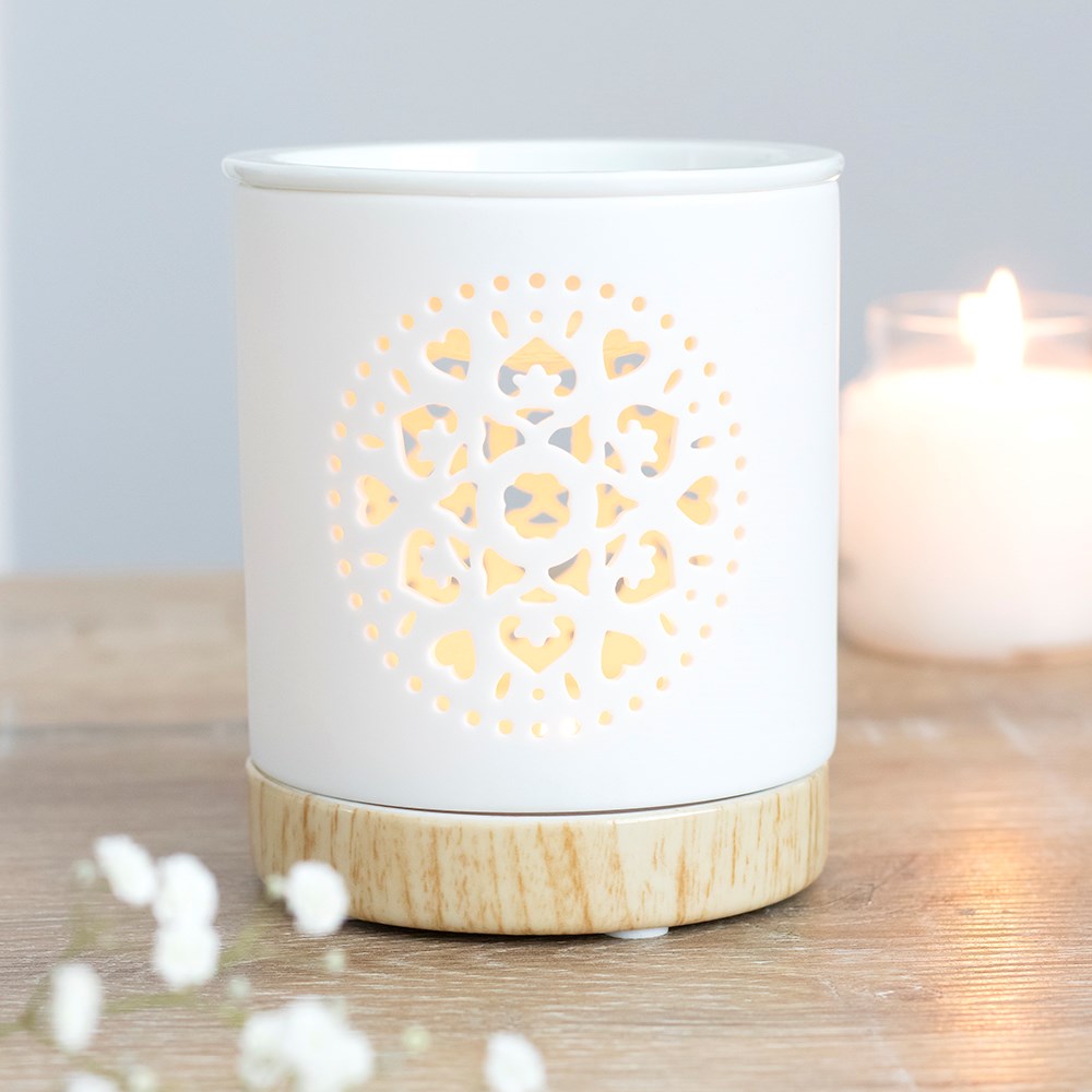 White cylindrical ceramic wax warmer with mandala cutout glowing on a light wood-effect base, beside a blurred candle and small white flowers.