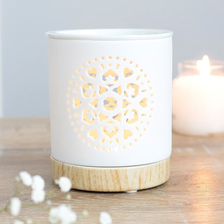White cylindrical ceramic wax warmer with mandala cutout glowing on a light wood-effect base, beside a blurred candle and small white flowers.