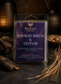 Clear plastic clamshell wax-melt pack with a deep purple label, upright on dark stone beside charred wood and dried grasses.